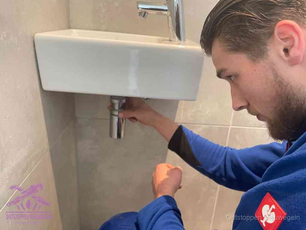 Een monteur draait met de hand de zwanenhalssifon onder een fonteinwasbak in de toiletruimte los om de bron van rioollucht in Nieuwegein te vinden